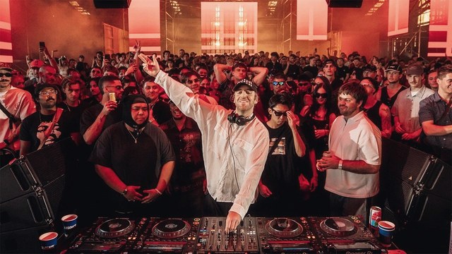 A DJ arms aloft behind the decks, surrounded on all sides by the crowd in a red-lit Boiler Room set