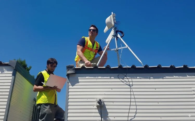 Matt Hall in hi-vis on a rooftop aligning a tripod-mounted antenna under a clear blue sky, a colleague below checking a laptop