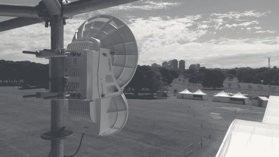 A point-to-point radio dish on scaffold overlooking the festival parkland site, graphite monochrome